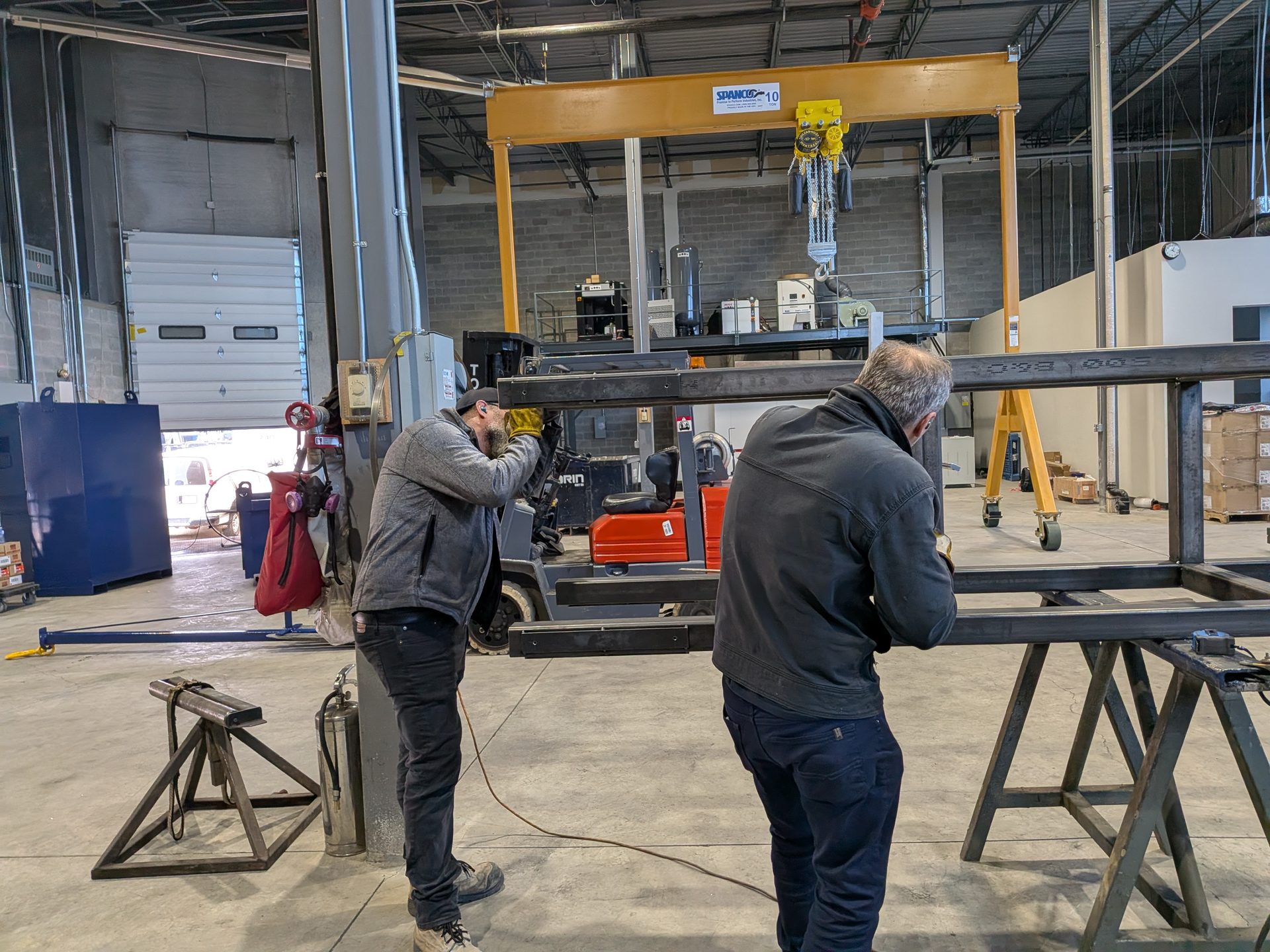 Two fabricators assembling a large steel frame beneath an overhead gantry crane